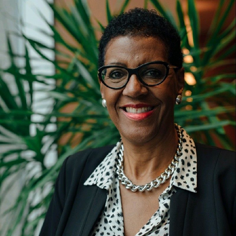 Deborah Simon wearing glasses and a necklace is smiling in front of a plant.