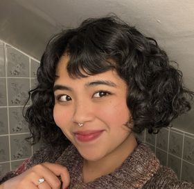 A woman wearing a denim jacket and a purple top is smiling in front of a body of water.
