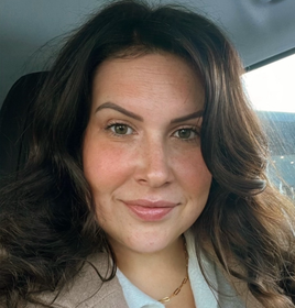 A woman wearing a denim jacket and a purple top is smiling in front of a body of water.