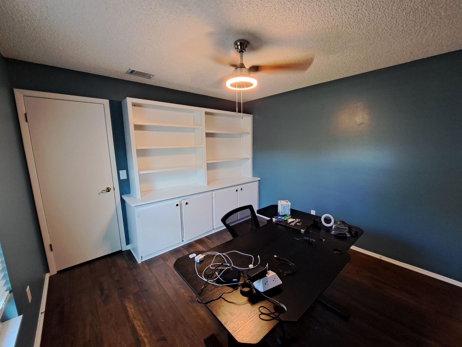 A room with a desk , chairs , shelves and a ceiling fan.