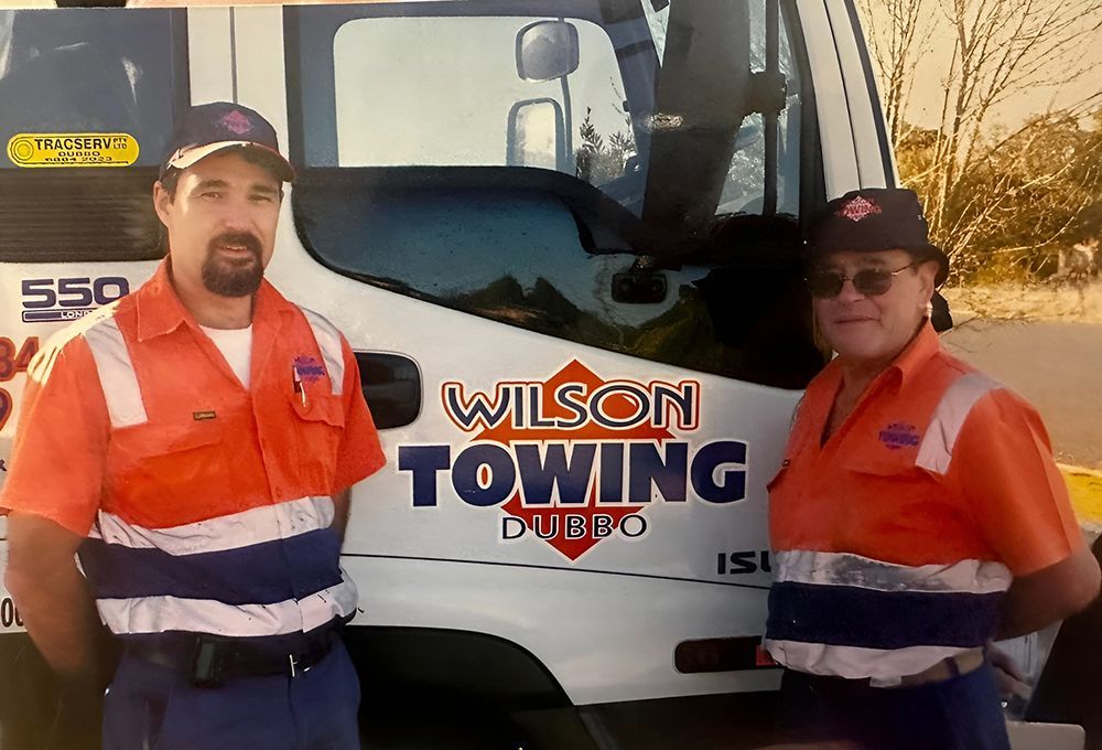 Two Men in Orange and Blue Uniforms Stand in Front of a Wilson Towing Truck — Tow in Dubbo, NSW