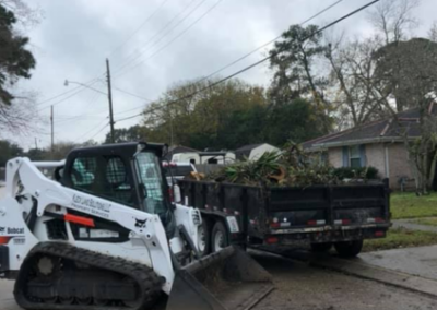 Bobcat skid-steer loader filling a trailer with tree branches on a street in front of a house.