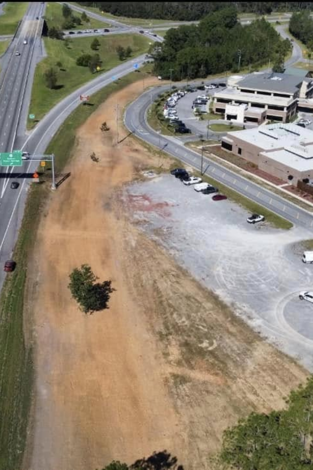 Aerial view of a bare dirt field alongside a highway and buildings, with some trees and a parking lot.