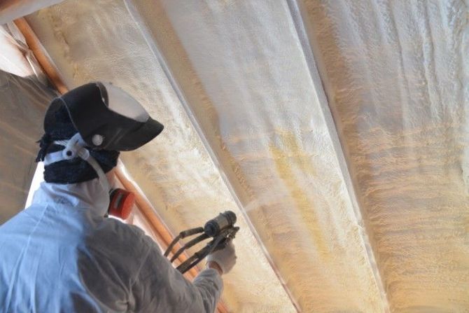 Worker spraying insulation onto attic ceiling with a spray gun while wearing protective gear