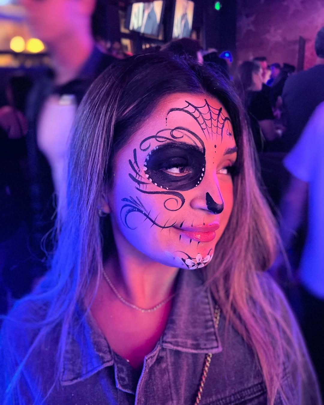 A woman with sugar skull makeup on her face is standing in a dark room.