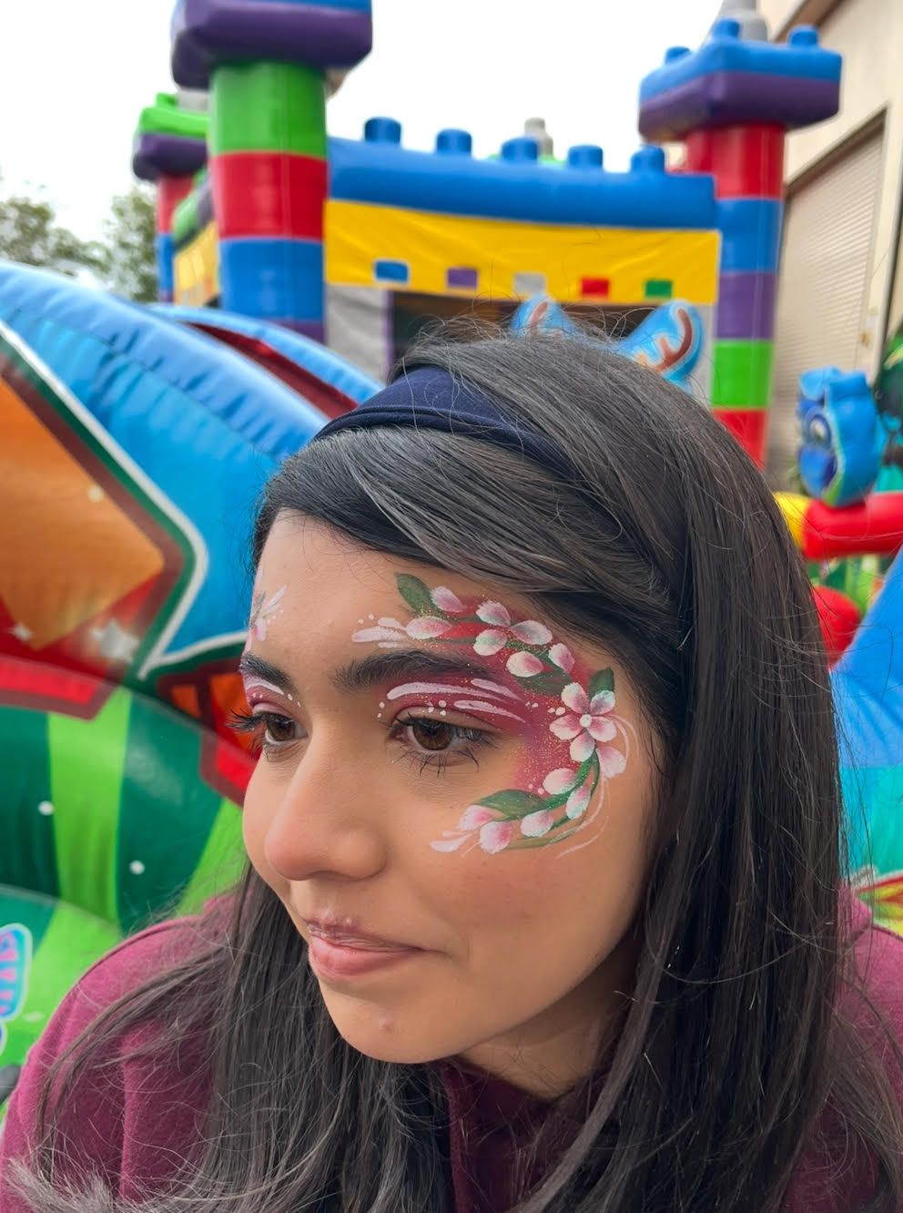 A woman with flowers painted on her face is standing in front of a bouncy house.