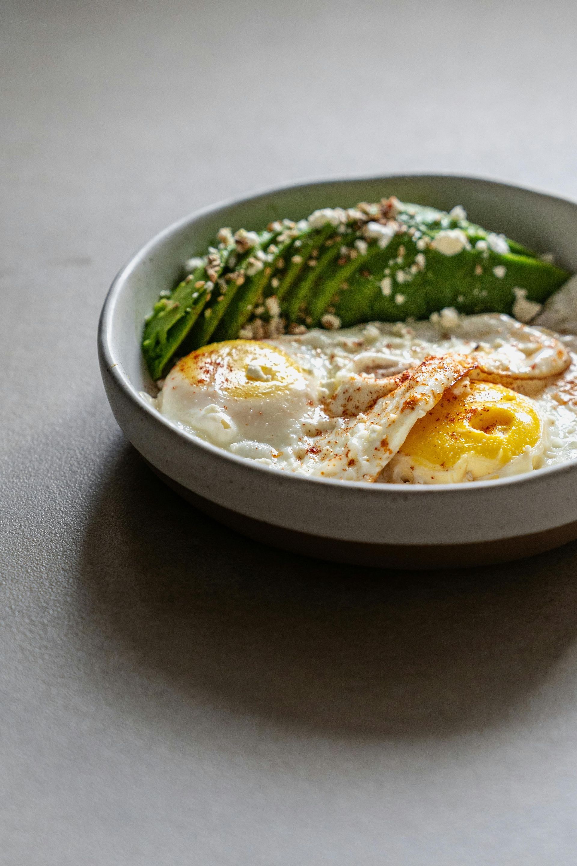 Breakfast bowl with sliced avocado, two fried eggs, and sprinkled topping.