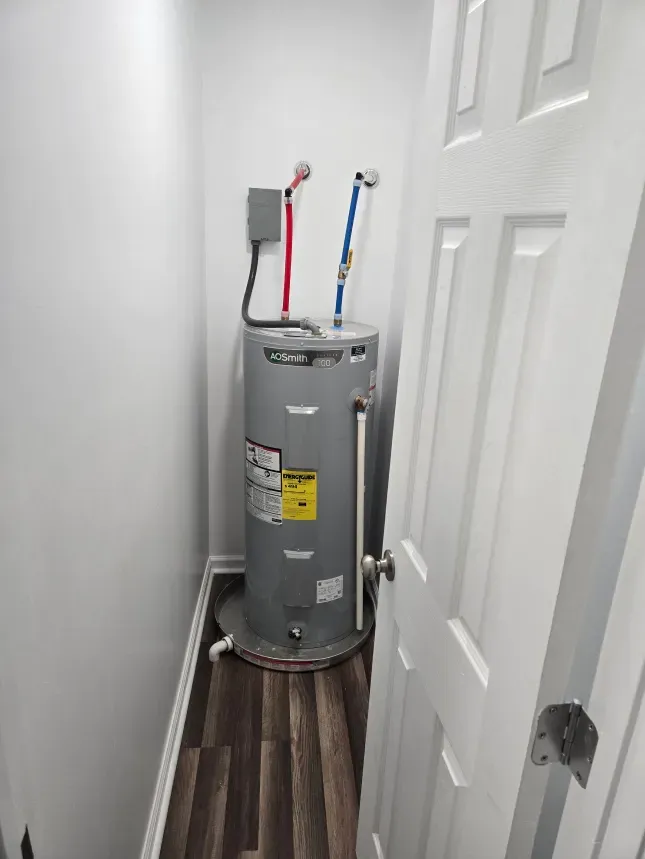 A water heater is sitting in a corner of a room next to a door.