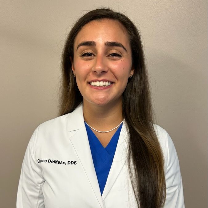 A woman in a white lab coat and blue scrubs is smiling for the camera.