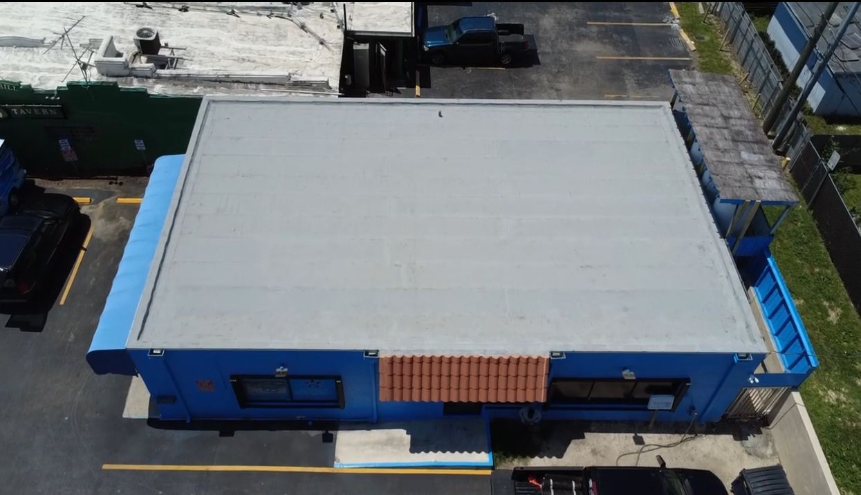 Overhead view of a blue building with a gray flat roof. Brown trim above a window, parking lot and fence.