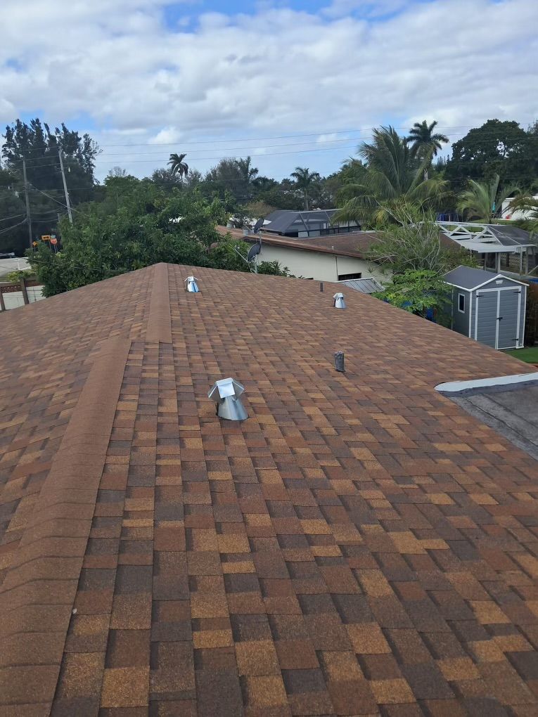 Brown shingled roof with vents, surrounded by trees, houses, and blue sky with clouds.