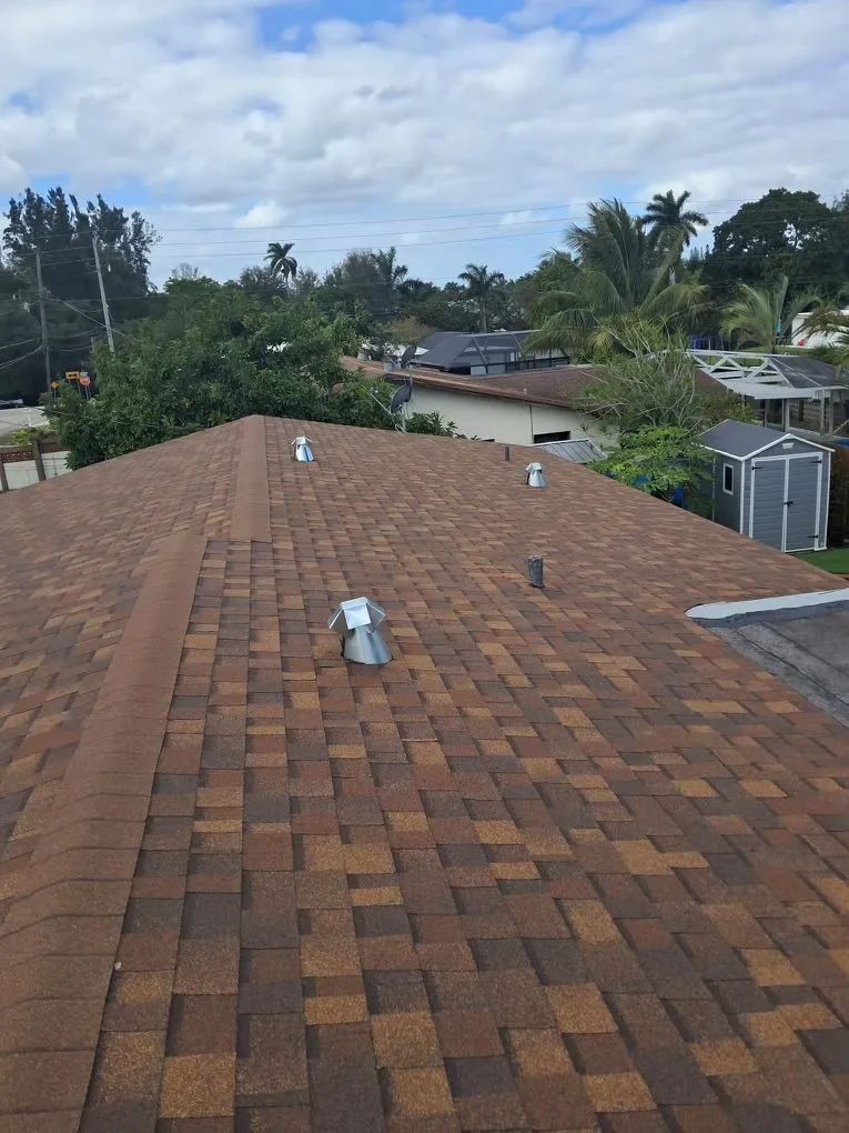 Brown shingled roof with vents. Blue sky with clouds and trees in the background.