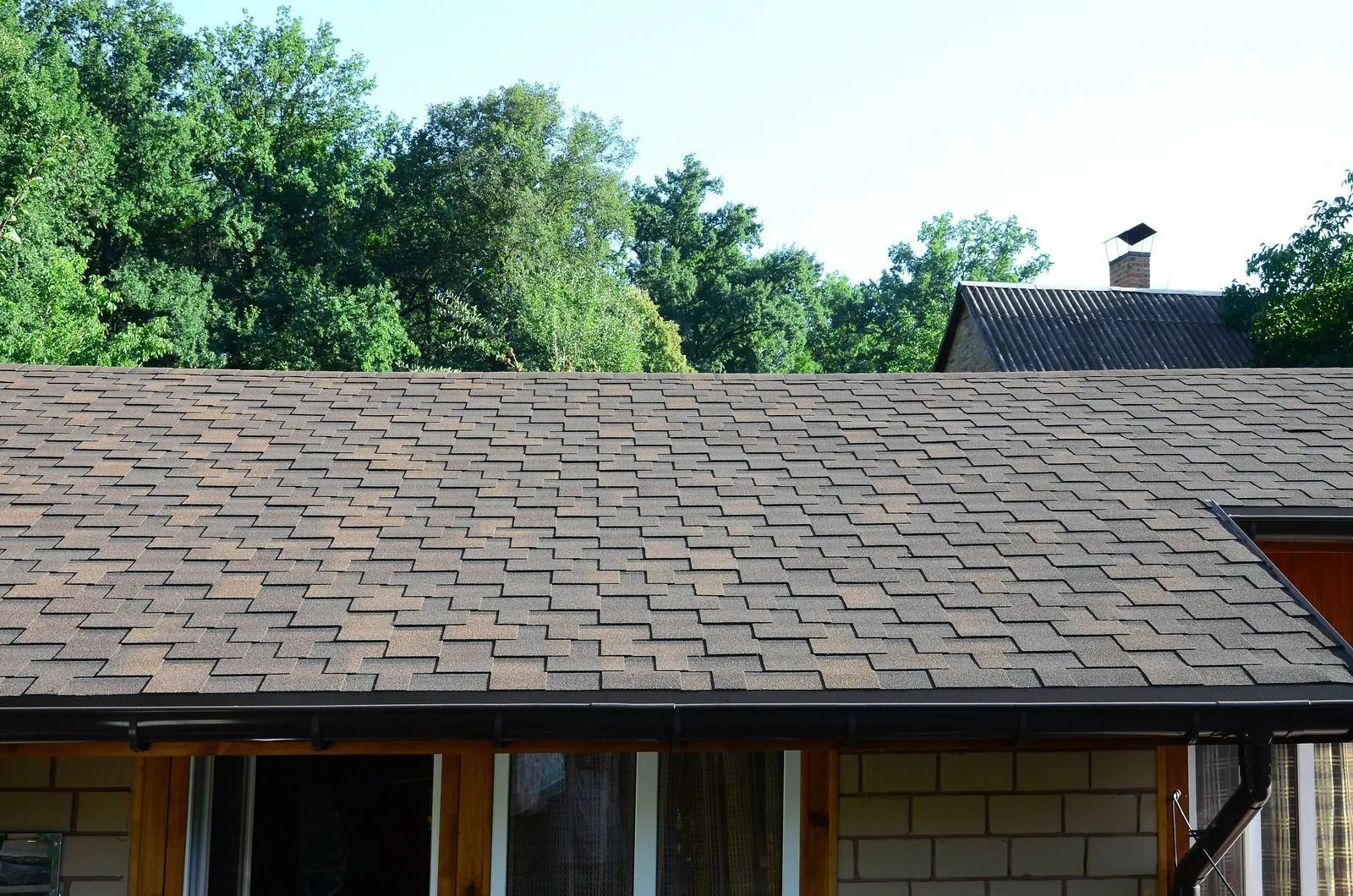Brown shingled roof with dark gutters; trees in background and part of another roof visible.