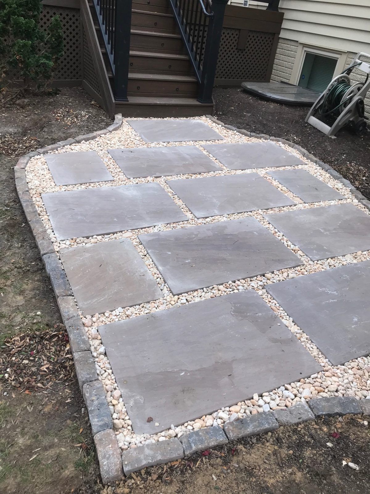 A new flagstone patio with gray rectangular stones, light gravel borders, and a stone edge, located at the base of stairs.