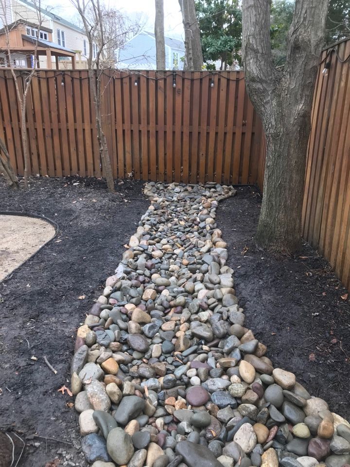A backyard area featuring a rock-filled drainage swale leading toward a wooden privacy fence near a large tree.