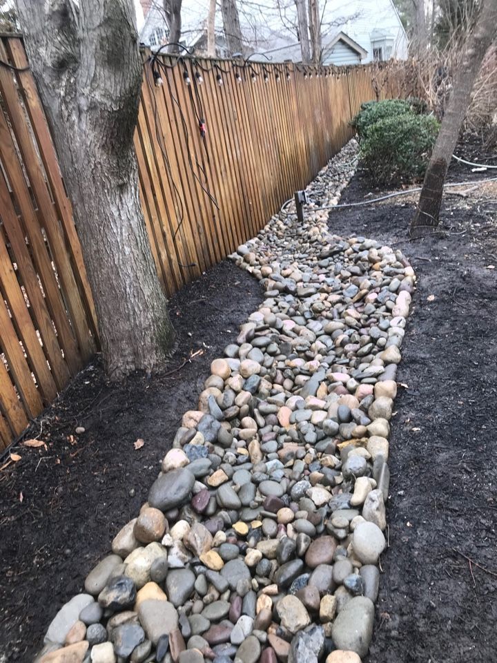 A rock-filled drainage ditch runs along a wooden fence next to a bare tree and dark soil in a yard.