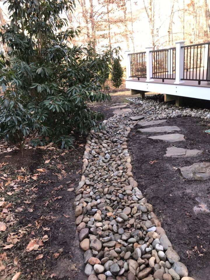 A stone path curves through a yard alongside a large shrub and an elevated wooden deck in a wooded backyard setting.