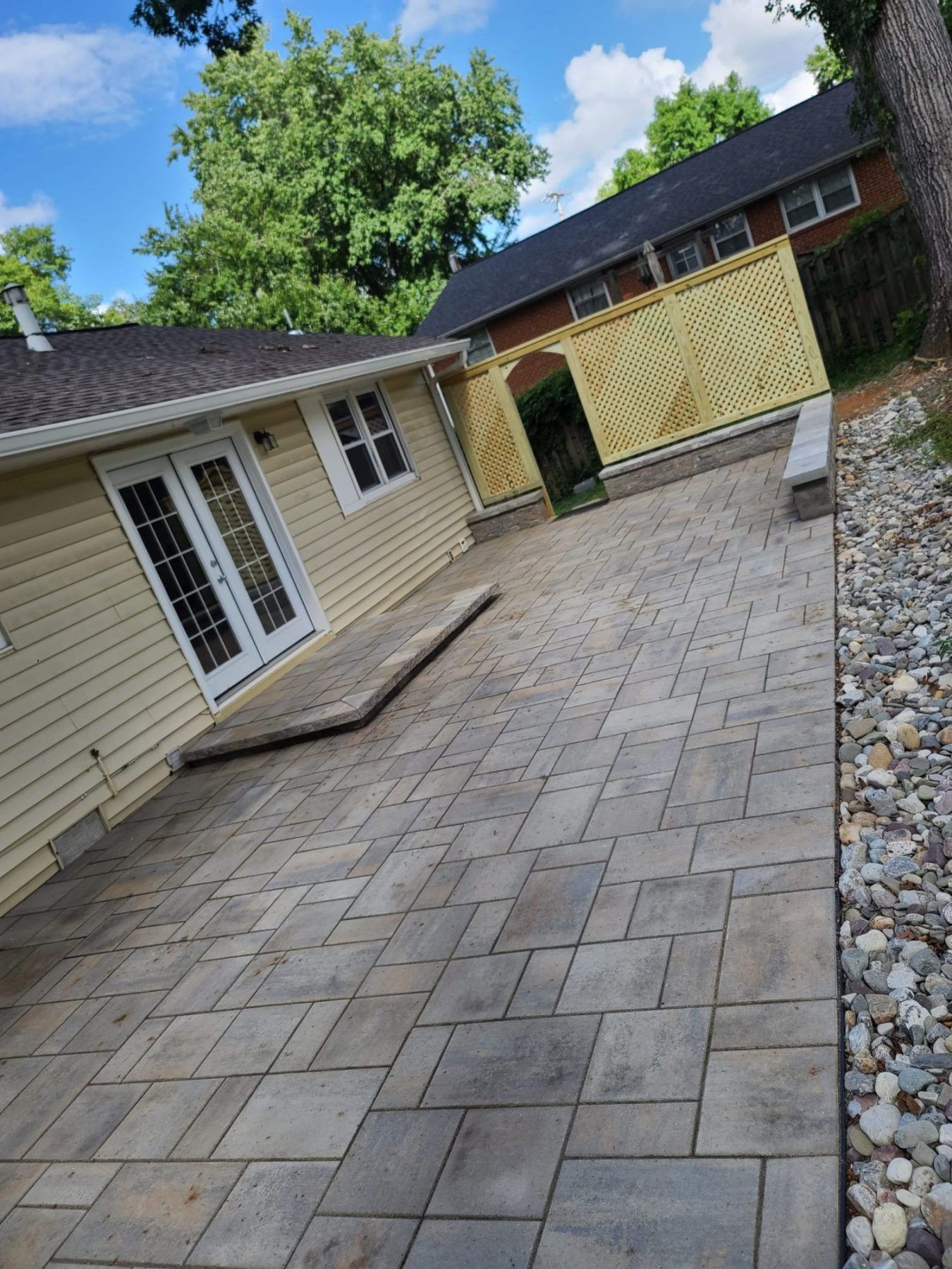 A paved stone patio alongside a light yellow house with French doors, featuring a lattice fence and stone borders.