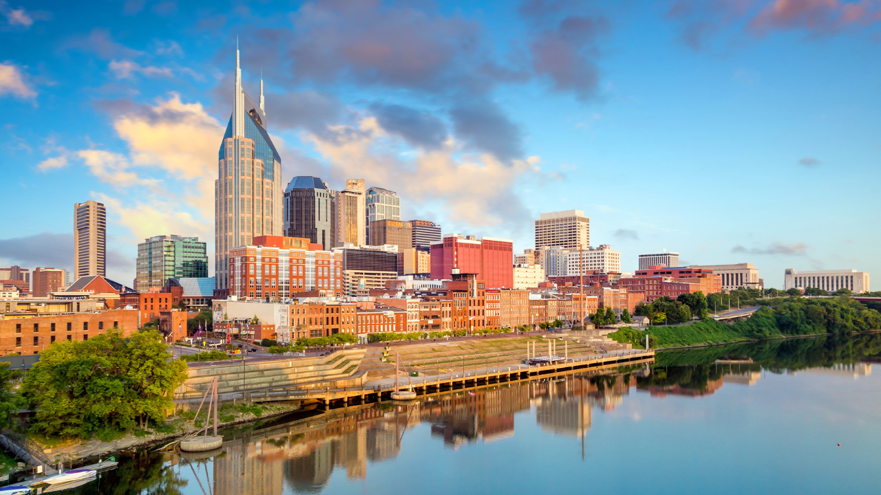 Skyline of downtown Nashville at sunset, with riverfront buildings and calm water reflections.