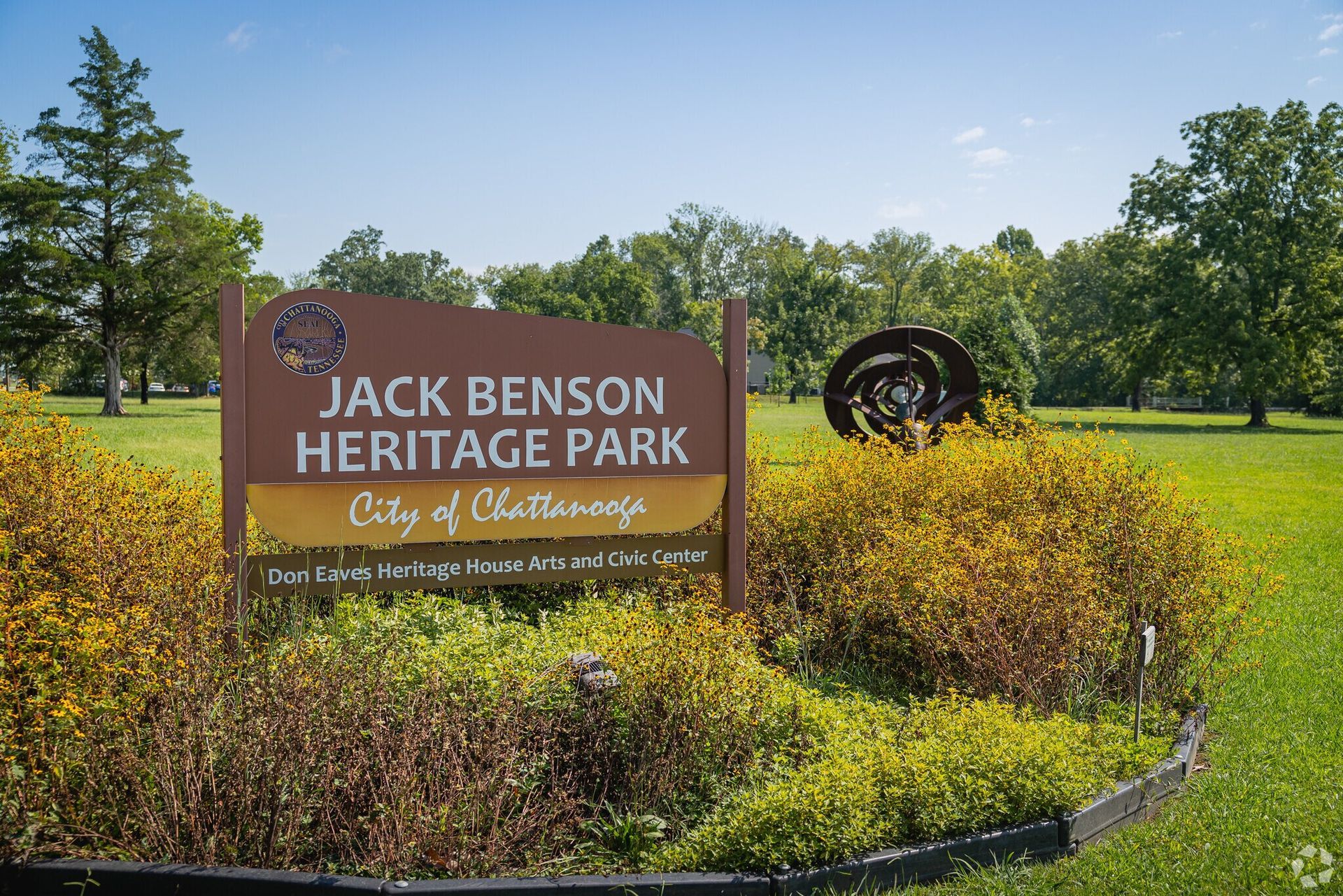 Jack Benson Heritage Park sign surrounded by shrubs and trees on a sunny day