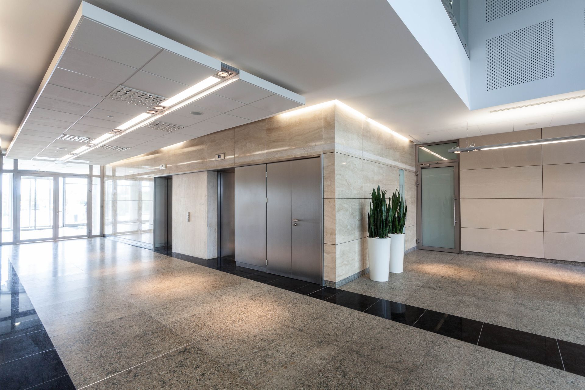 Modern office lobby with stainless steel elevators, polished stone floor, and potted plant by glass doors