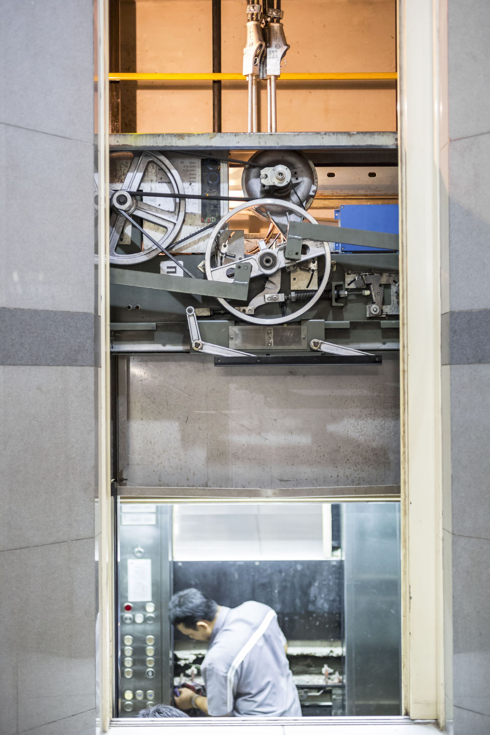 Elevator shaft with exposed machinery above a worker in a gray hoodie inside the cabin