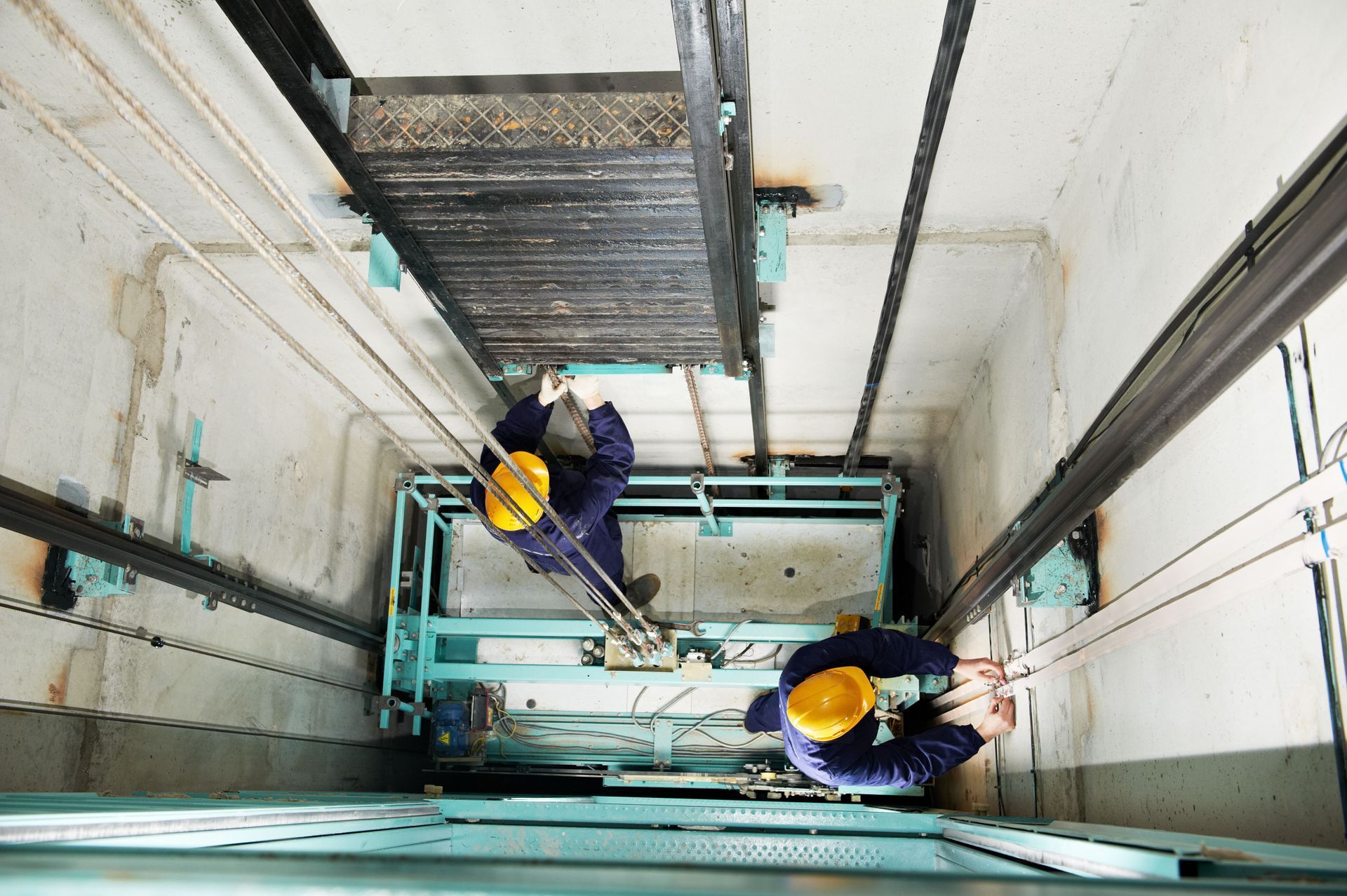 Two workers in yellow hard hats inside a vertical industrial shaft, viewed from above.