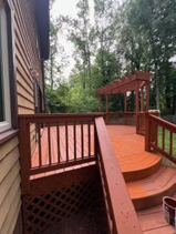 Wooden deck and staircase with red railings beside a house, surrounded by trees.