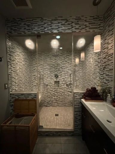 A modern bathroom featuring a walk-in glass shower with textured stone walls, a wooden hamper, and a sink vanity.