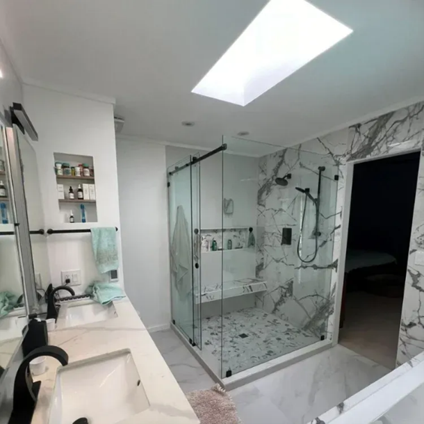 Modern bathroom with marble shower, glass door, and skylight. Vanity and open doorway visible.
