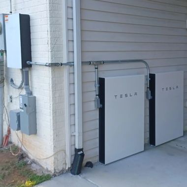 Two Tesla Powerwall batteries mounted outside a house beside electrical panels and conduit.