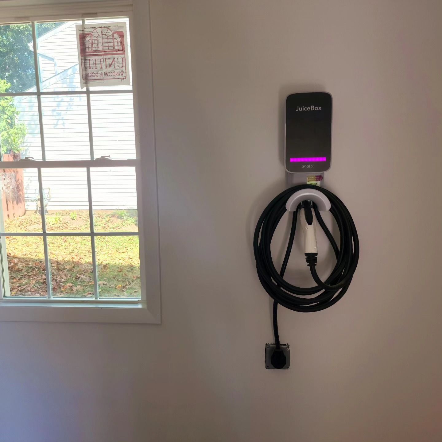 Wall-mounted EV charger with coiled cable and purple light beside a window.