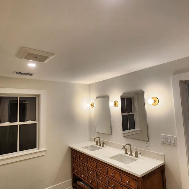 Bright bathroom with double sinks, wood vanity, mirrors, sconces, and a window on the left.