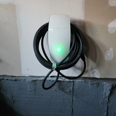 White wall-mounted device with a glowing green light and coiled black cable against an unfinished wall.