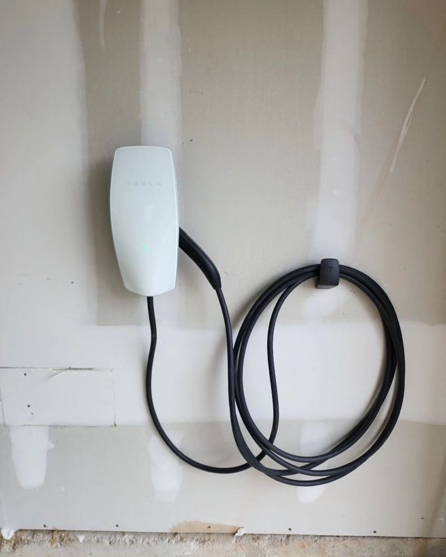 White wall-mounted device with a coiled black cable hanging beside it on a plain unfinished wall