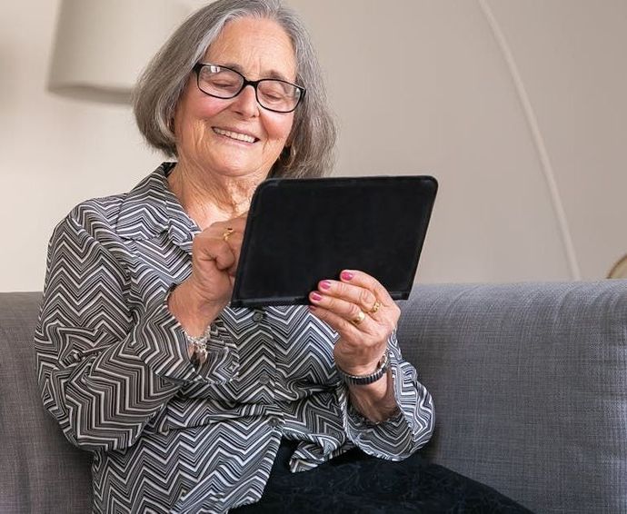 An elderly woman is sitting on a couch using a tablet computer.