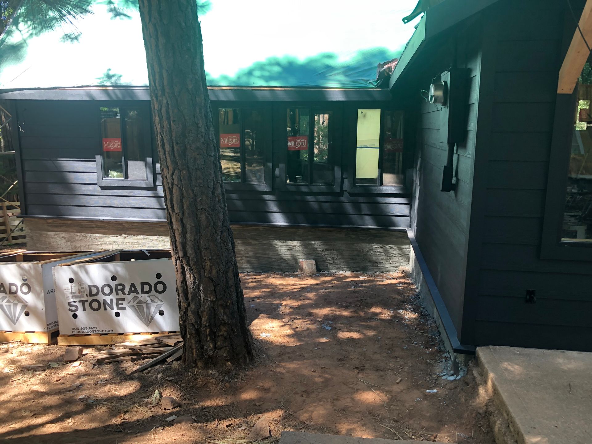 A dark-painted house exterior under construction, featuring stone cladding at the base and Eldorado Stone packaging nearby.