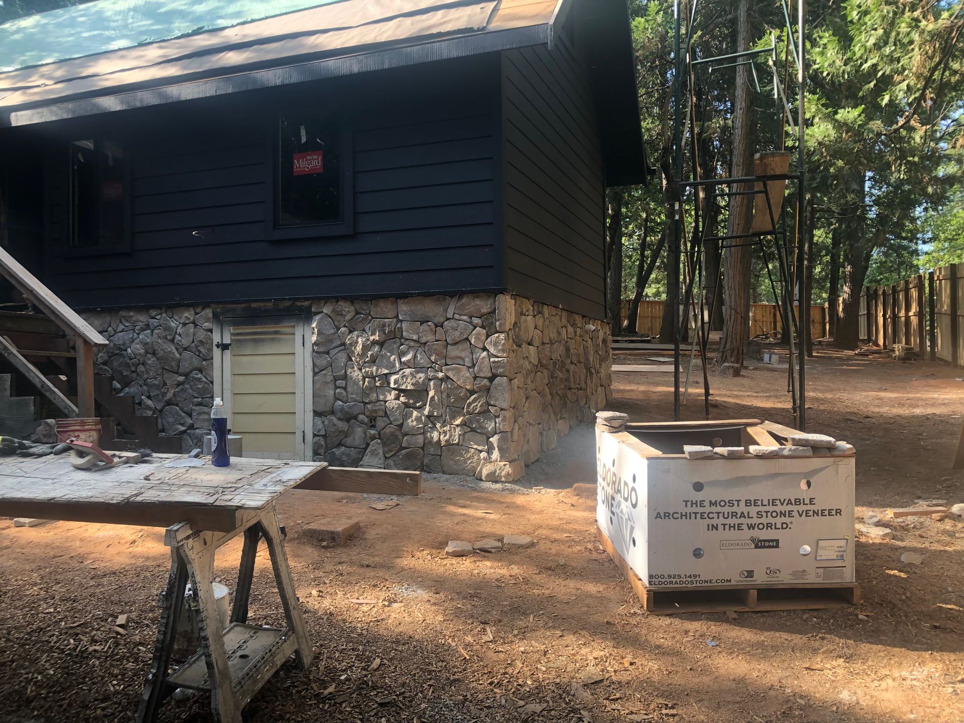 A black cabin with a stone foundation under construction, with a work table and construction supplies in the yard.