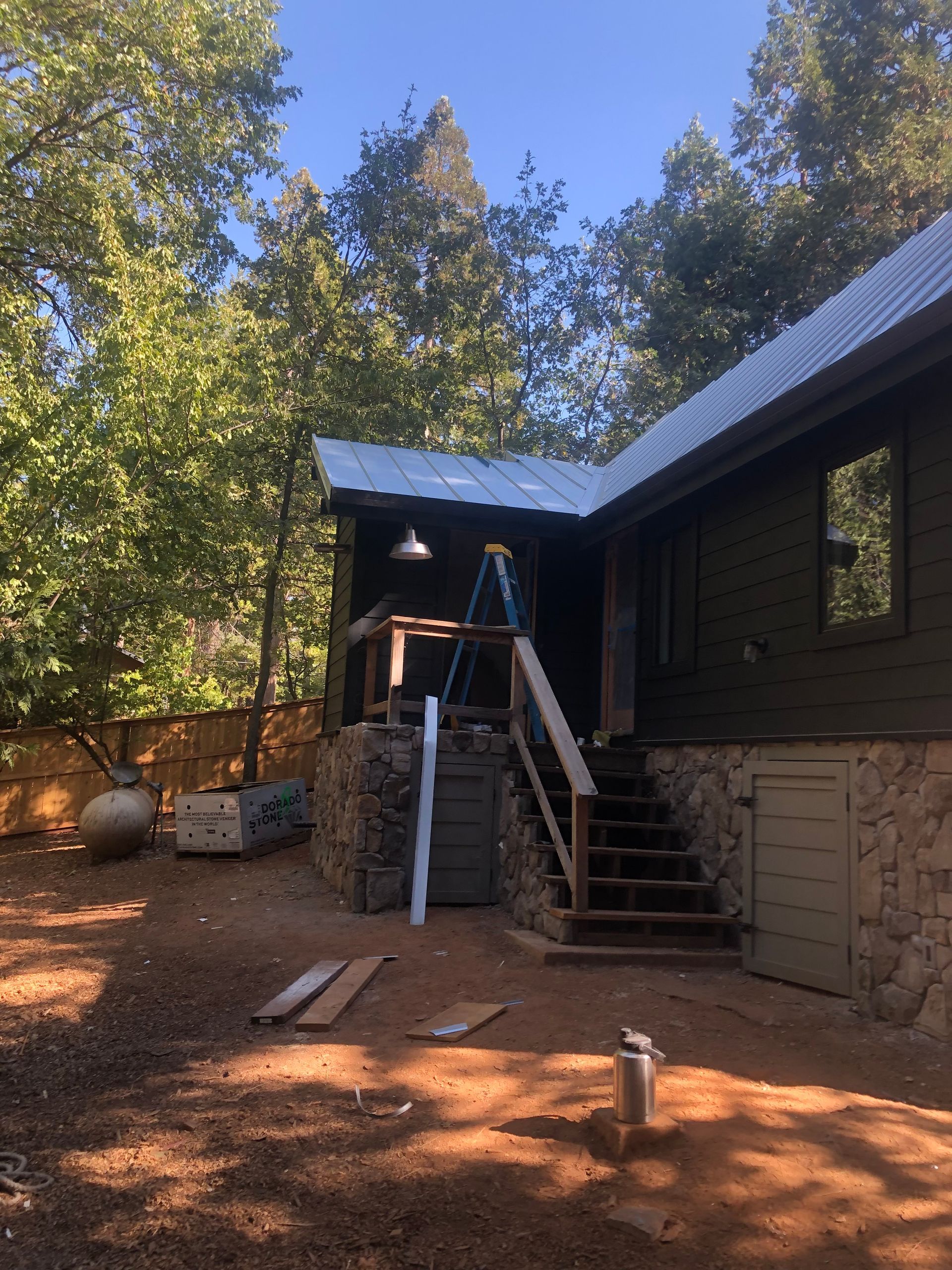 A wooden house with a stone foundation and a white metal roof, featuring a staircase leading to a porch under renovation.