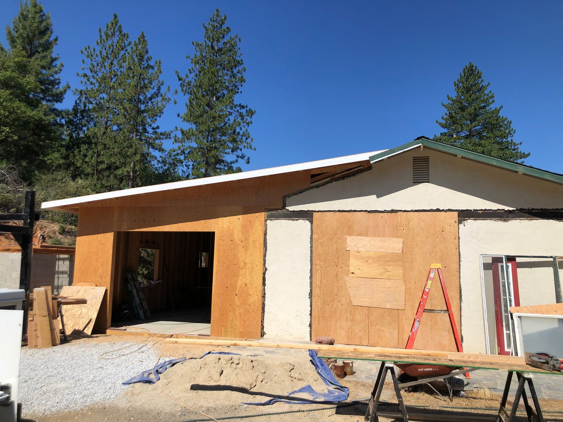 A construction site featuring an unfinished house with plywood walls, a garage opening, and a red ladder under a blue sky.