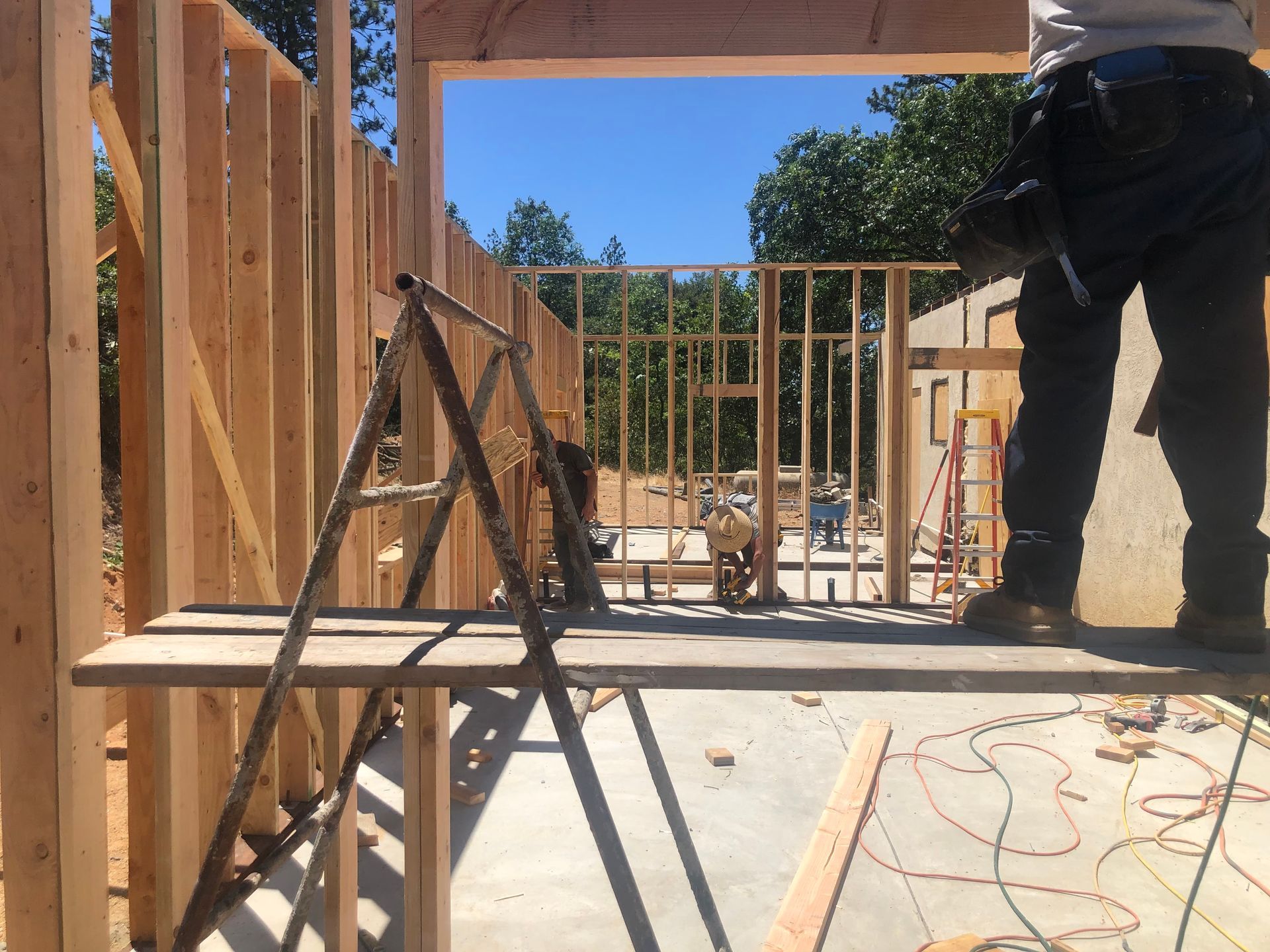 A worker stands on wooden scaffolding in an unfinished building, with wooden wall framing visible in the background.
