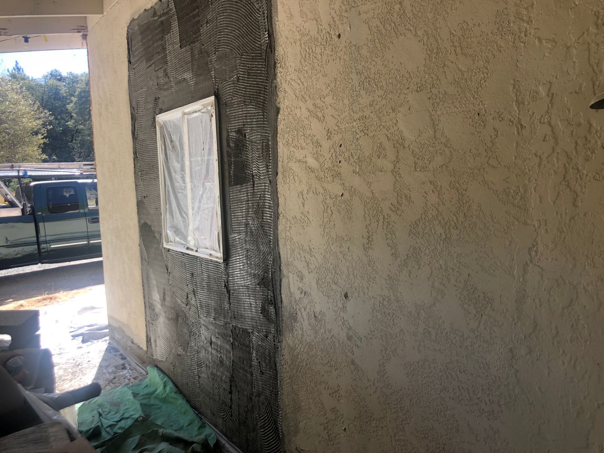A wall under construction with a patch of fresh gray mortar and a masked window next to an existing textured tan wall.