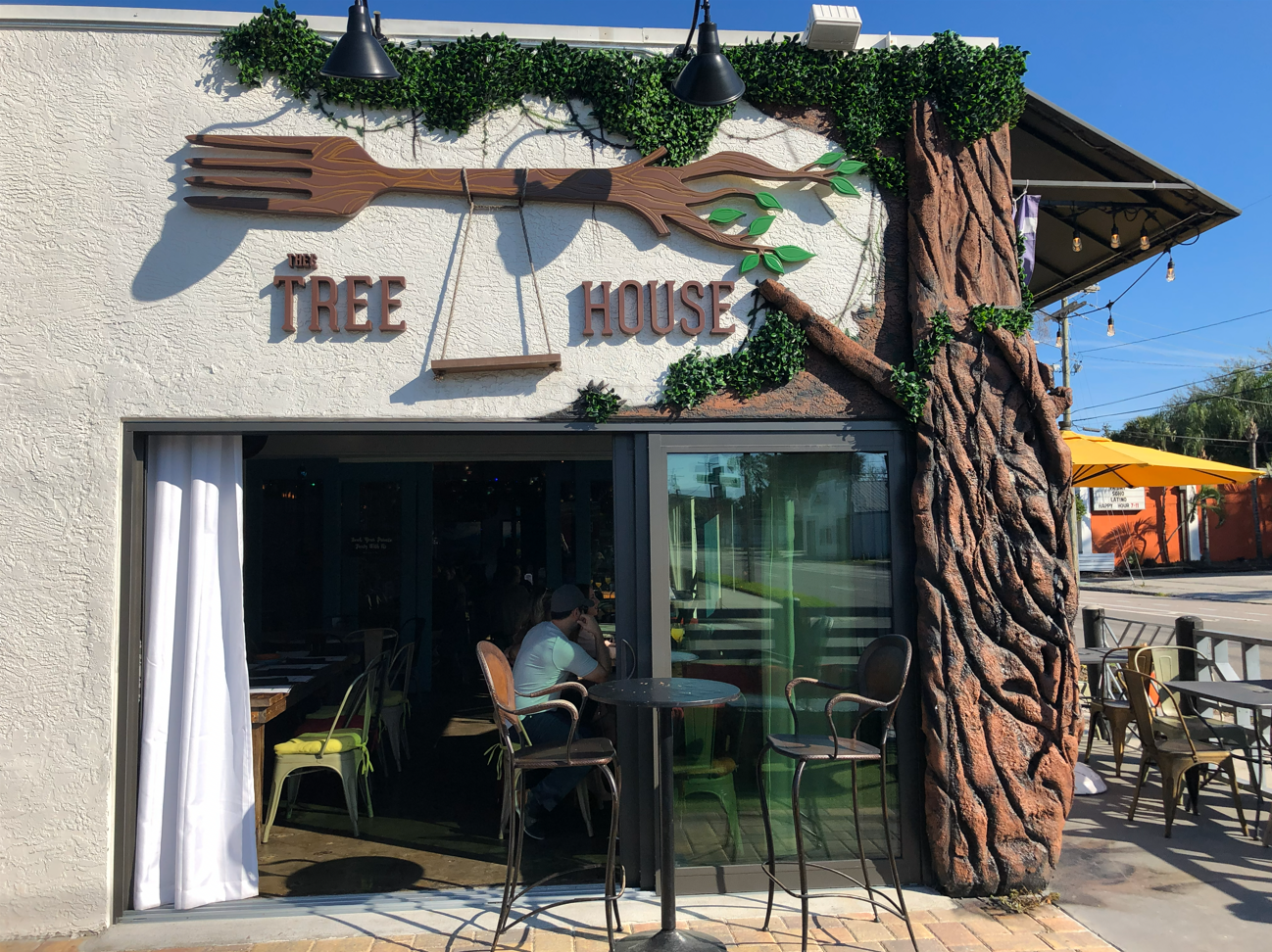 Entrance of Tree House restaurant with a fork sign, tree trunk facade, and outdoor seating.