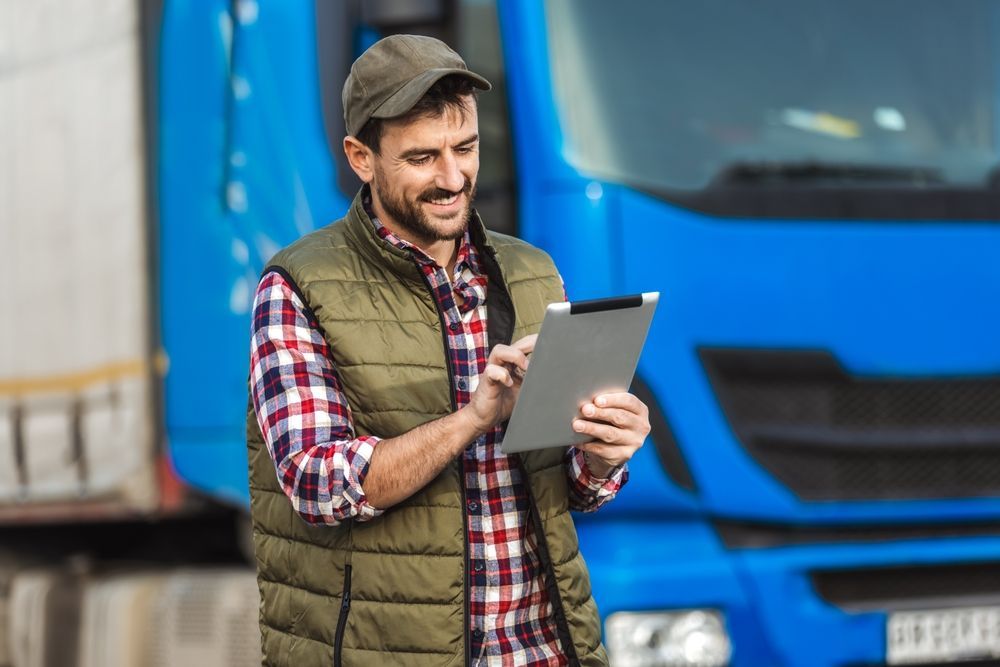 Truck driver in a plaid shirt and vest, using a tablet, standing in front of a blue truck.