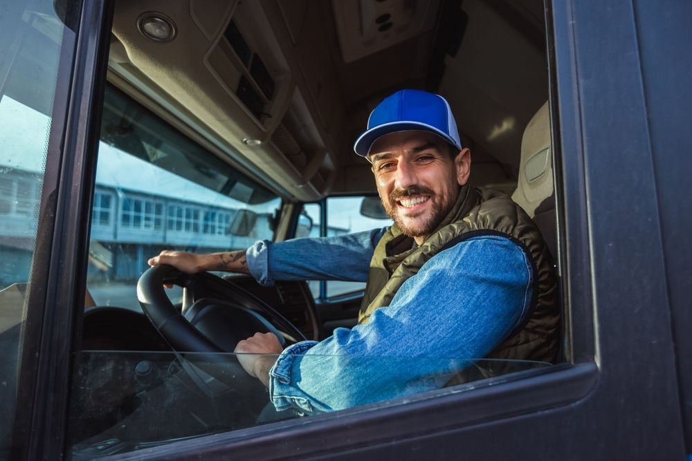 Smiling truck driver in a blue cap and denim shirt, sitting behind the wheel.