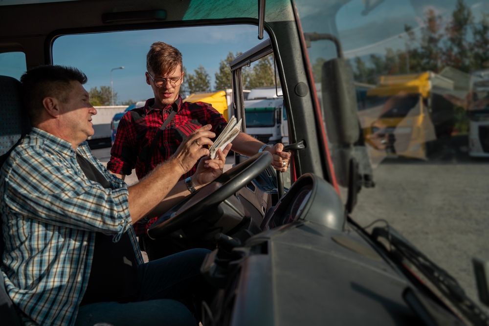 Two people in a truck cab, one driving and one looking at a map in a farmyard