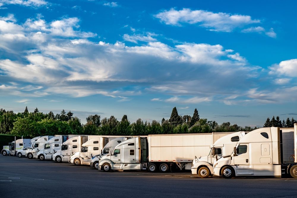 Row of white semi trucks parked in a lot under a blue sky with scattered clouds