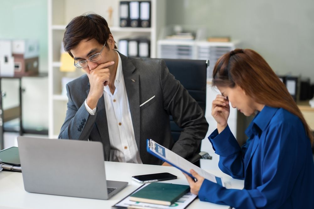 Two professionals sit at a desk, looking stressed while reviewing documents on a laptop and a clipboard in an office.