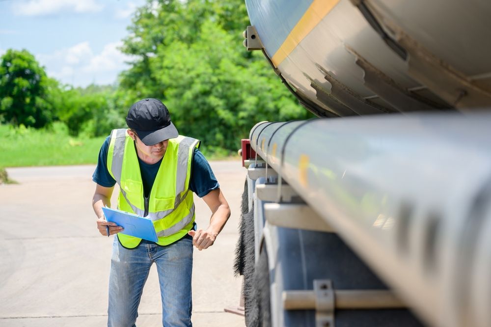 Man in safety vest inspecting a tanker truck, holding a clipboard outdoors.