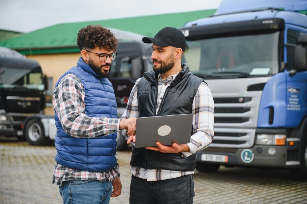 Two truck drivers discussing route optimization and fleet management on a laptop, standing next to commercial trucks